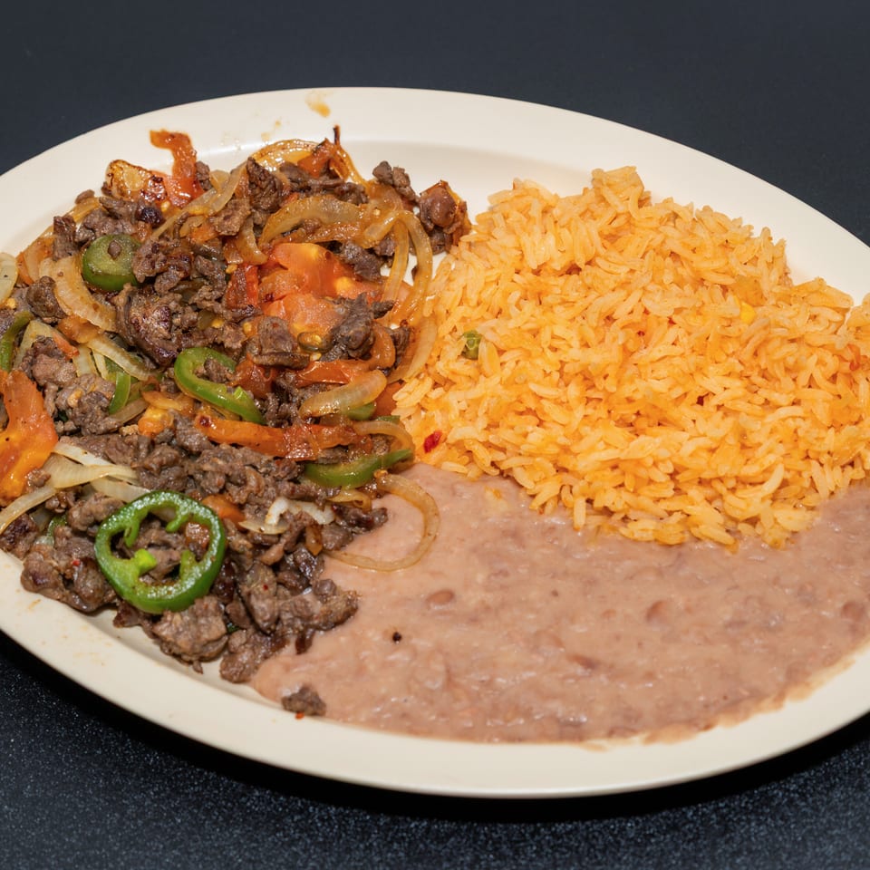 Plate of seasoned beef with peppers, onions, and tomatoes, served with Mexican rice and refried beans.
