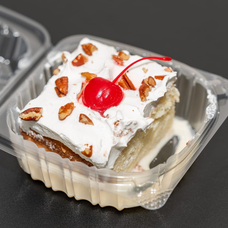 Slice of layered cake topped with whipped cream, chopped pecans, and a red cherry in a clear takeout container.