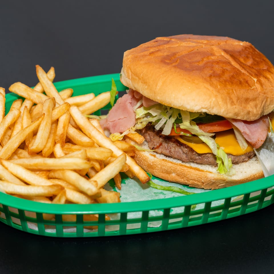 Cheeseburger with ham, lettuce, tomato, pickles, and onions served with golden fries in a green basket.