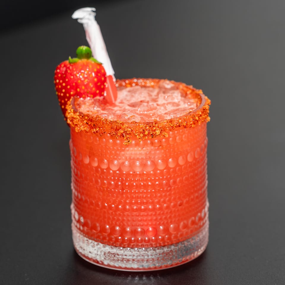 Red cocktail in a textured glass with a chili-rimmed edge, strawberry garnish, and white straw against a dark background.
