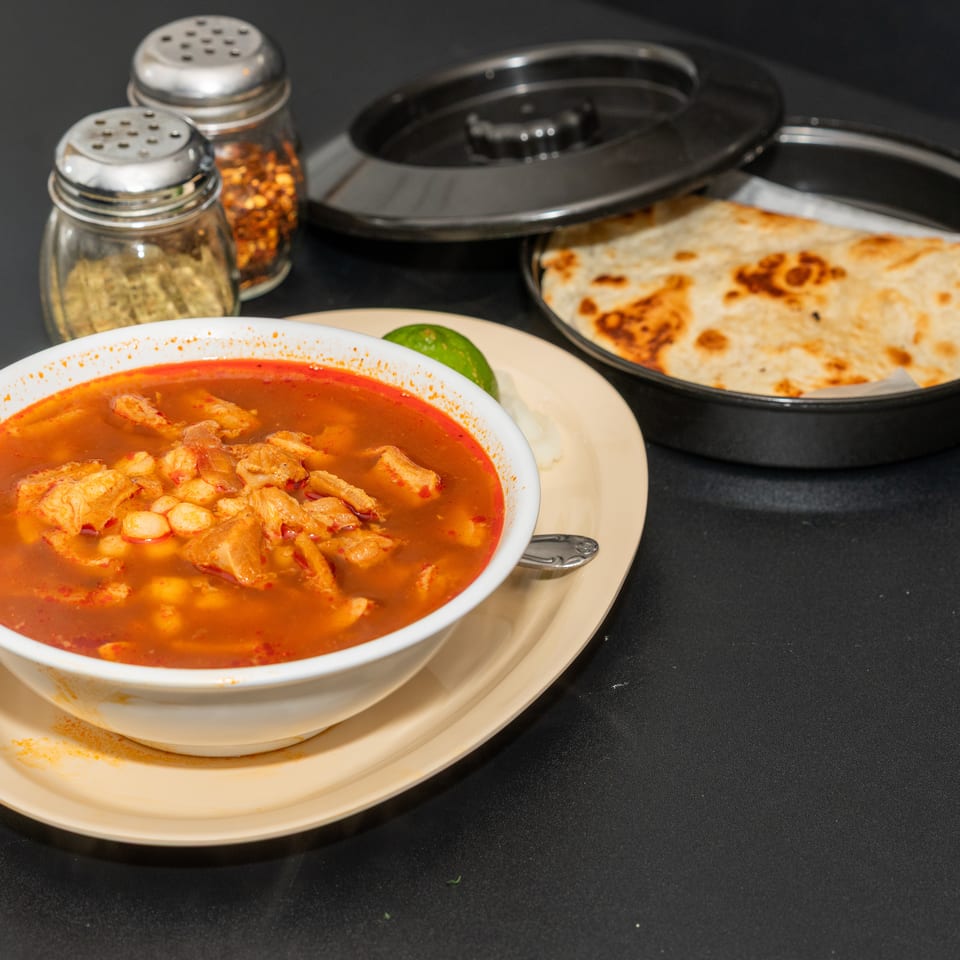 Bowl of spicy red soup with meat beside toasted flatbread, lime, and spice shakers on a restaurant table.